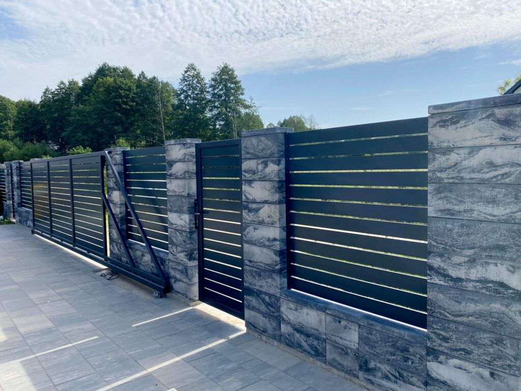 Tamsiai pilka melsva betoninė tvora su horizontaliais tamsiai pilkais metaliniais segmentais ir vartais, kurių dalis atidaryta, melsvai pilkos spalvos.