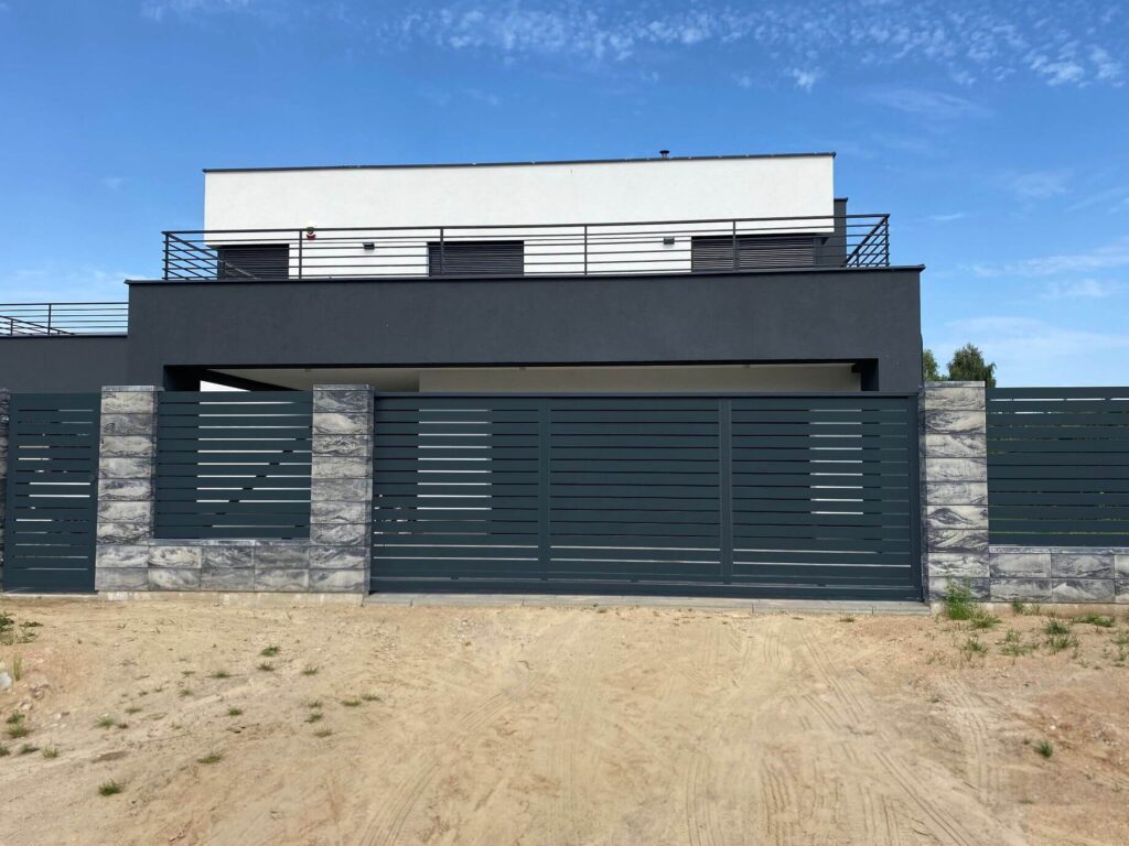 Moderni tamsiai pilka metalinė horizontalių segmentų tvora su šviesiai margomis betono kolonomis prie šiuolaikinio balto ir juodo namo.