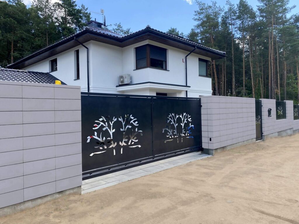 Tamsiai pilkos metalinės vartų ir tvoros segmentai su išpjaustytais medžių motyvais, šviesių betoninių tvoros kolonų fone, prie balto namo miško pakraštyje.