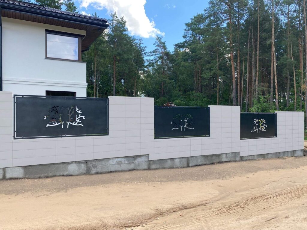 Šviesių betono tvoros segmentai su tamsiai pilkais metaliniais įstatytais langeliais, dekoruotais medžių motyvais, prie balto dviejų aukštų namo miško fone.