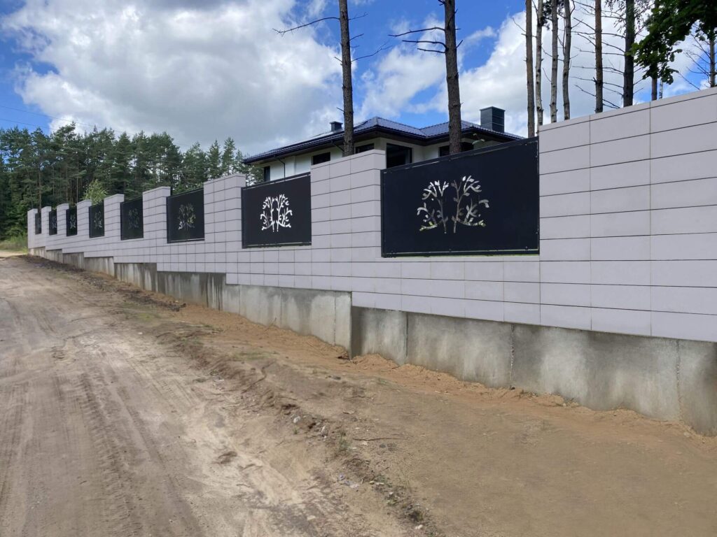 Šviesių betono tvoros segmentai su juodais metaliniais įdėklais, dekoruotais stilizuotais augalų motyvais, fone matomas namas ir pušynas, prie tvoros žemė su smėliu.