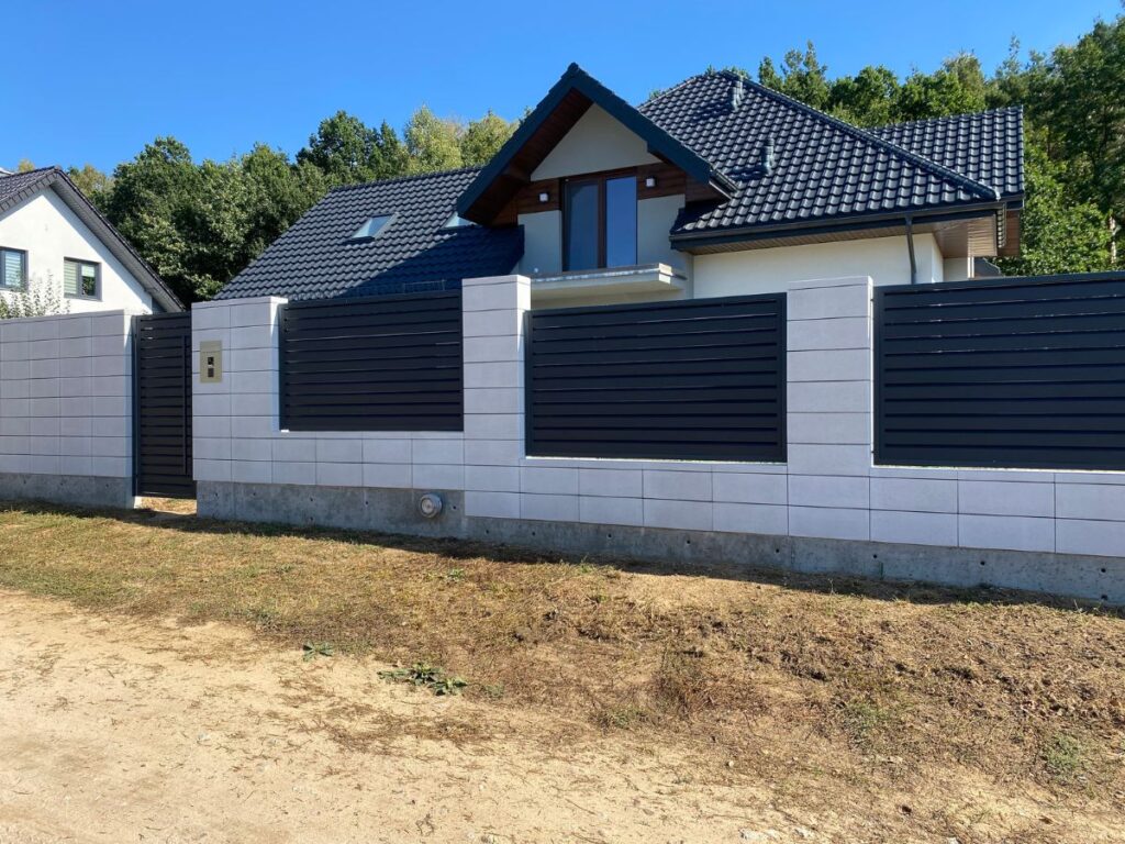 Šviesiai pilki betoniniai tvoros stulpai su horizontaliais tamsiai pilkais metaliniais tvoros segmentais ir vartais šalia gyvenamojo namo.