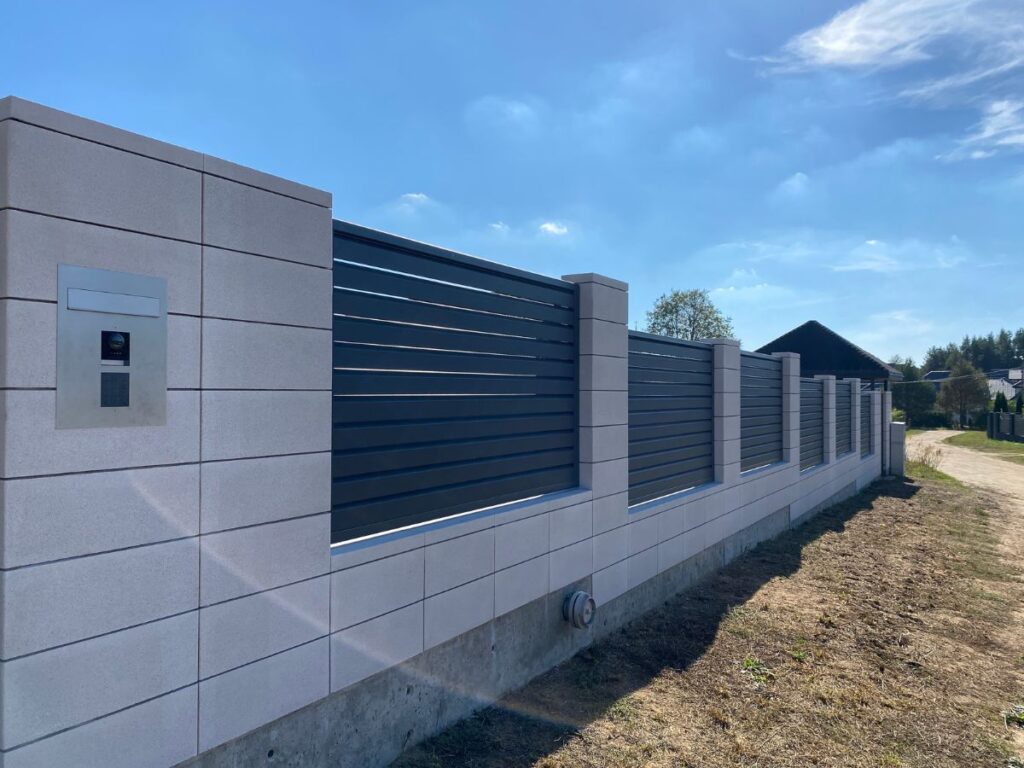 Šviesiai pilki betoniniai tvoros stulpai ir tvora su horizontaliomis tamsiai pilkomis metalinėmis juostomis, šviesiai pilka betoninė tvora su tamsiai pilkais metaliniais vartais ir įėjimo sistema.