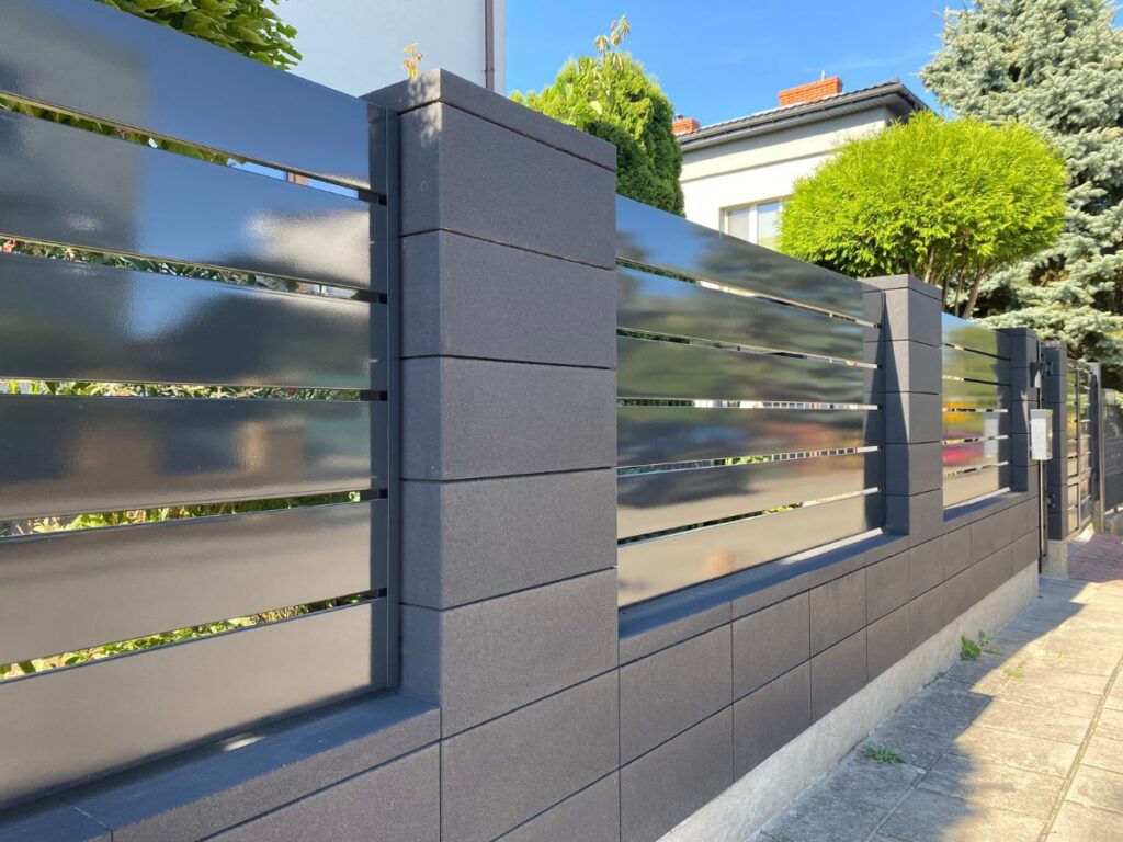 Tamsiai pilkos spalvos betoniniai tvoros stulpai ir apačioje blokeliai su blizgiais tamsiai pilkais metaliniais horizontaliais segmentais.