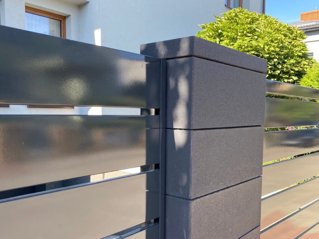 Tamsiai pilkas betoninis tvoros stulpas su horizontaliais blizgiais tamsiai pilkais metaliniais tvoros segmentais.