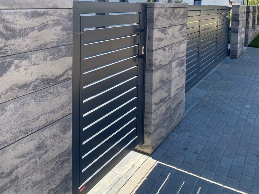 Tamsiai pilka metalinė pėsčiųjų vartų dalis su horizontaliomis juostelėmis, įmontuota į šviesiai pilką marmuro rašto tvoros stulpą, grindinys iš pilkų trinkelių.
