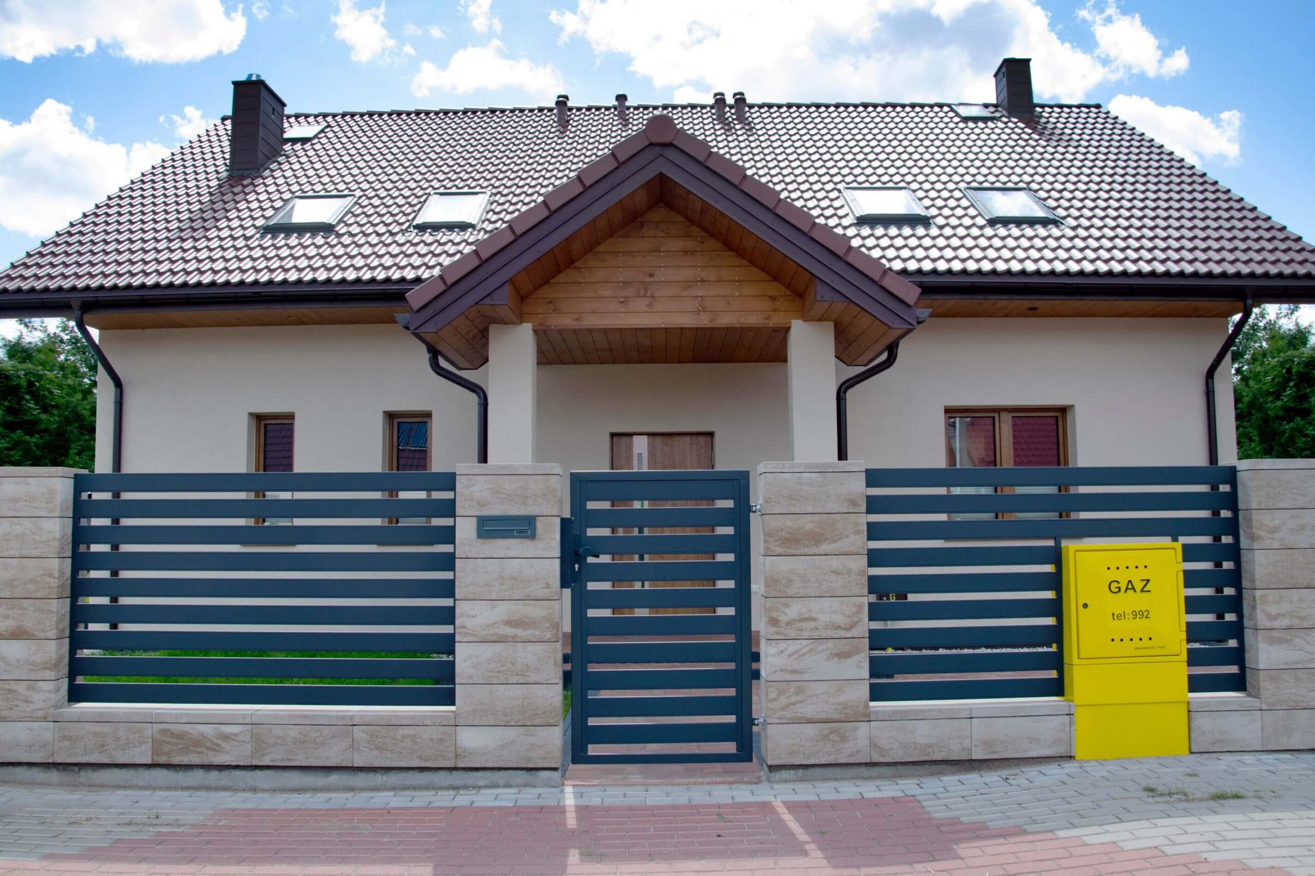 Tamsiai pilkos horizontaliai metalinės tvoros grotelės su vartais tarp šviesių akmens stulpų, šalia tvoros ryškiai geltonas dujų dėžės skyrius, už tvoros matomas namas.