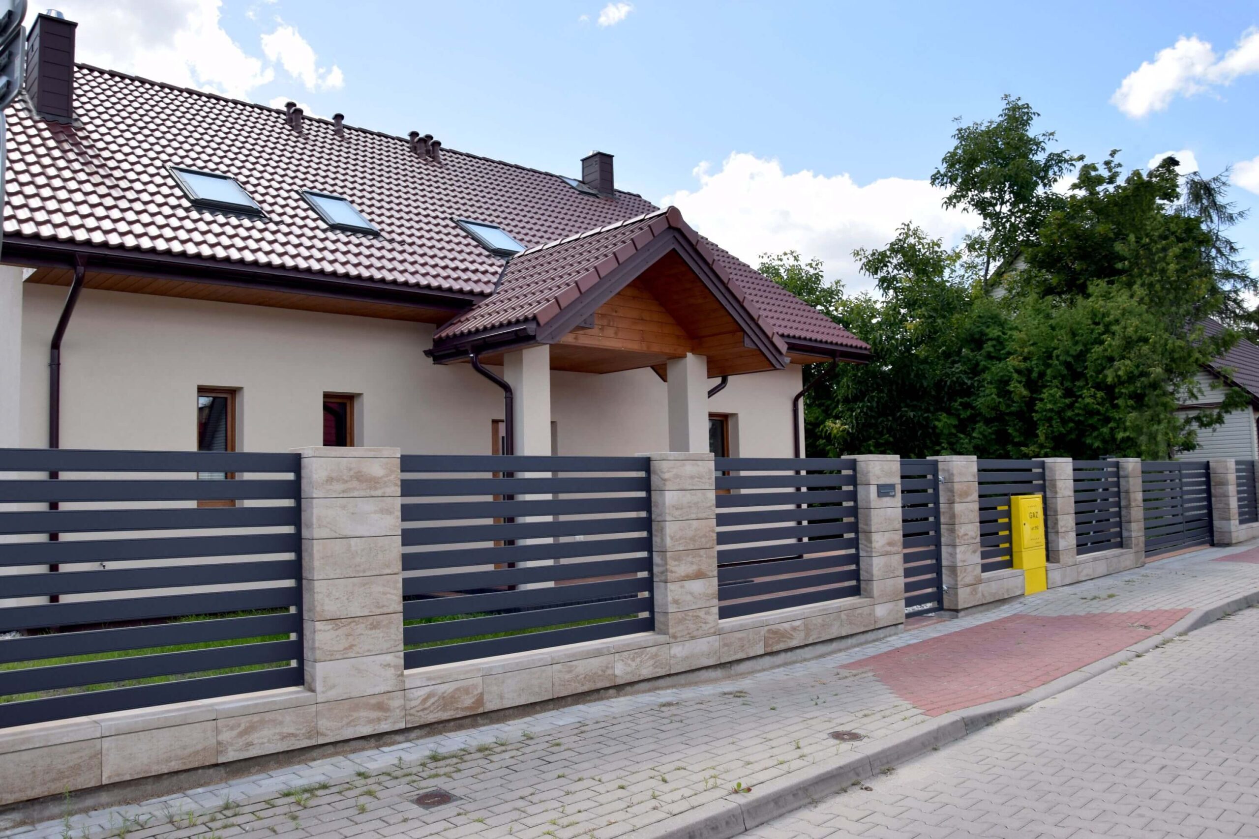 Tamsiai pilka horizontali metalinė tvora su metaline tvora vartais tarp šviesių akmens stulpų ir pamatų, gyvenamasis namas fone.