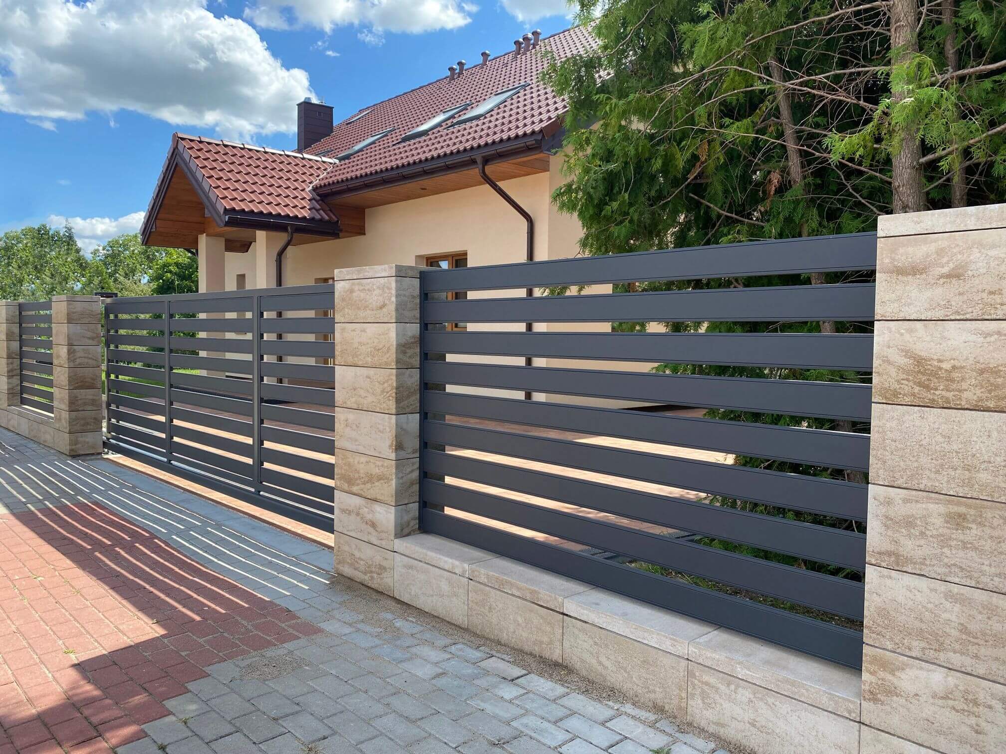 Tamsiai pilka horizontali metalinė tvora su vartais, įtvirtinta tarp šviesių akmens stulpų ir žemų tvorelės blokų, šalia raudonas plytelių takas.