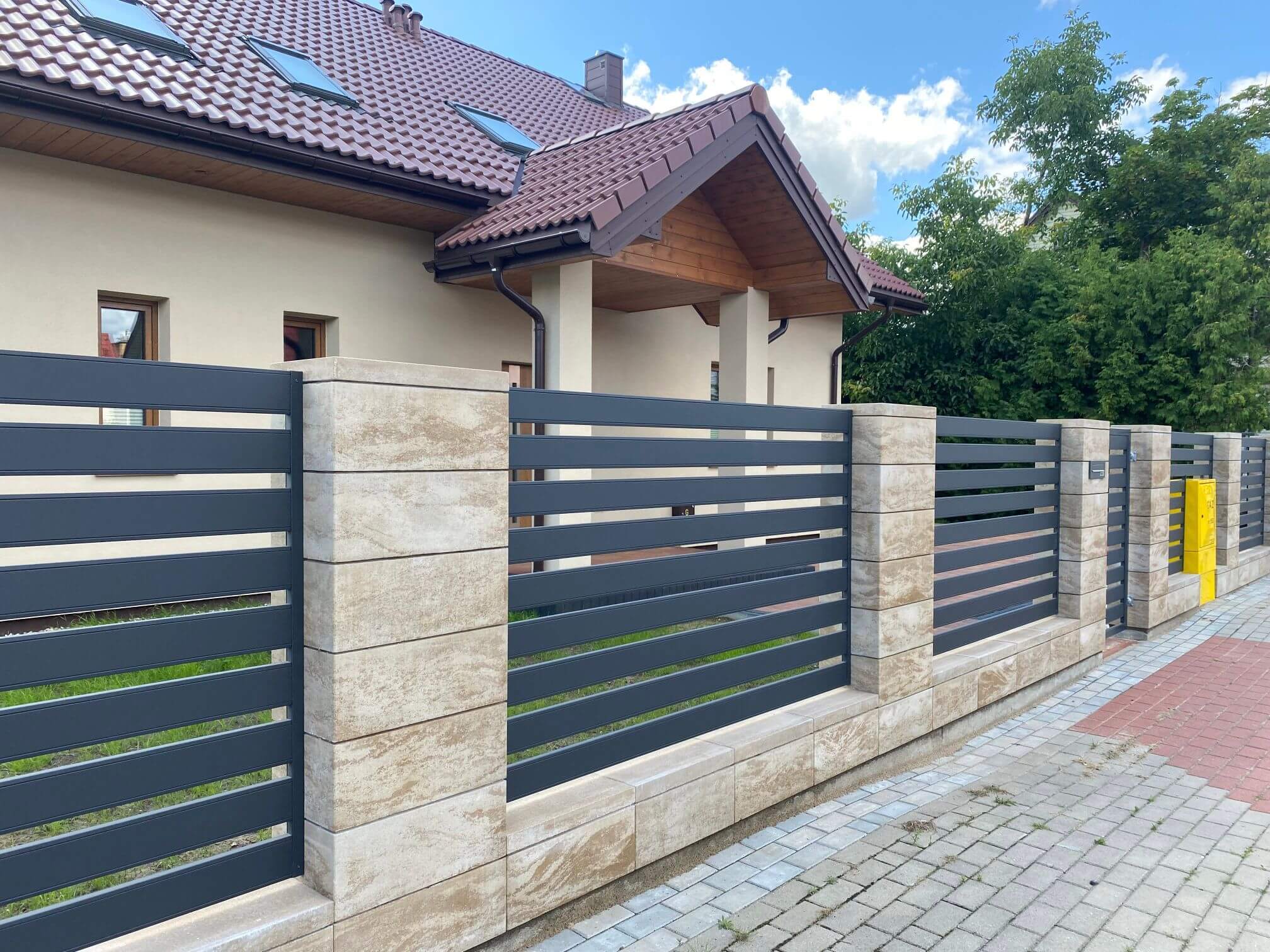 Tamsiai pilkos horizontaliai metalinės tvoros grotelės tarp šviesių akmens stulpų, už tvoros matoma namo dalis su langu.