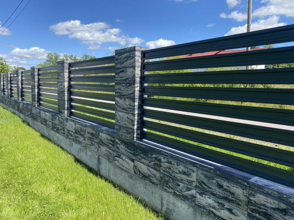 Pilka dekoratyvinė betoninė tvora su juodo metalo horizontaliomis juostomis, žalia veja prie tvoros